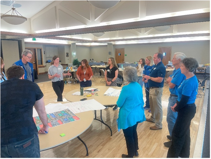 Group of people inside a large room with tables look at maps and plans, discussing among themselves