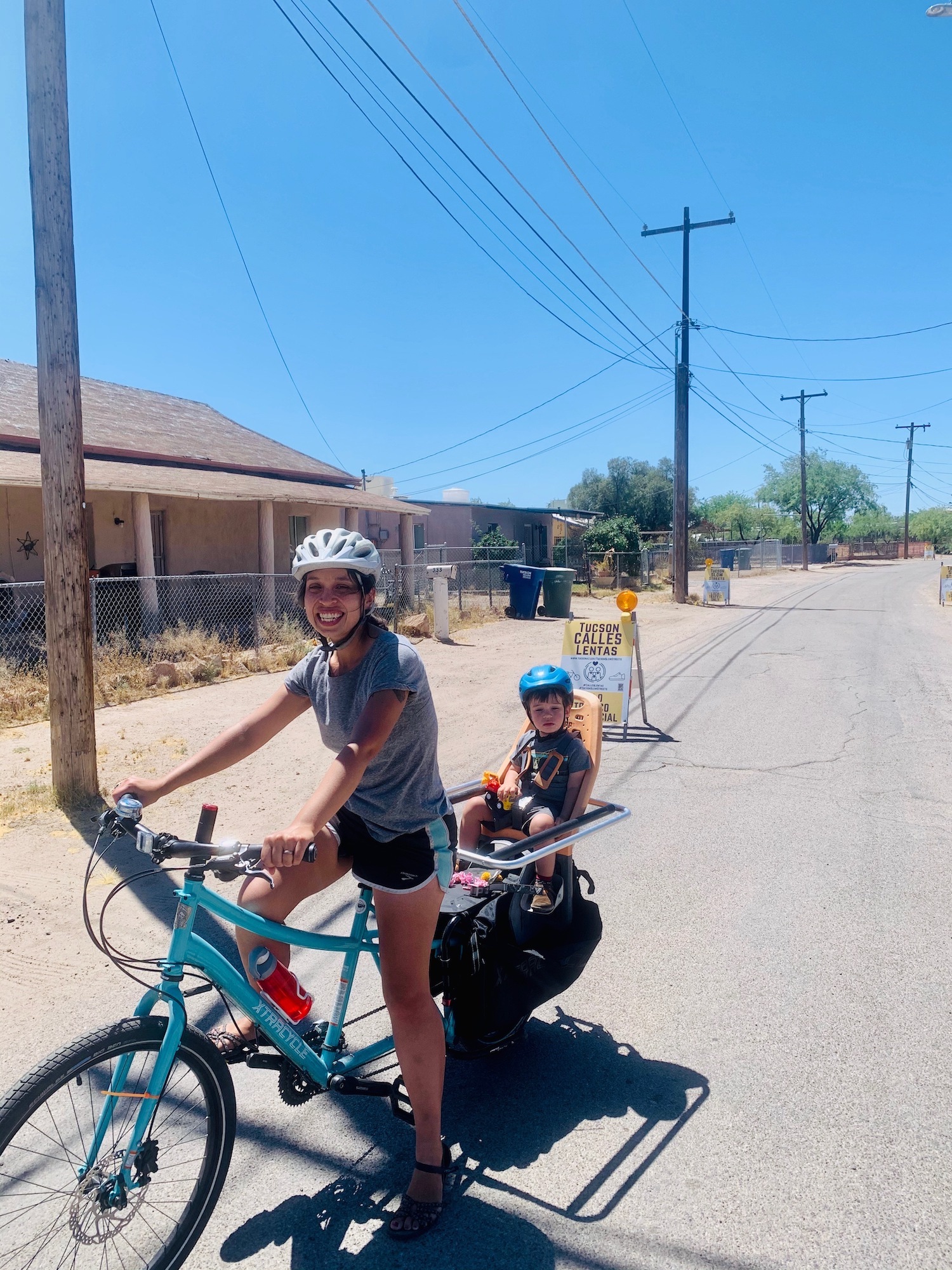 Woman on blue bike with one foot down to balance. Small child in a bike seat behind the woman. It is sunny and the bike is on a paved trail.