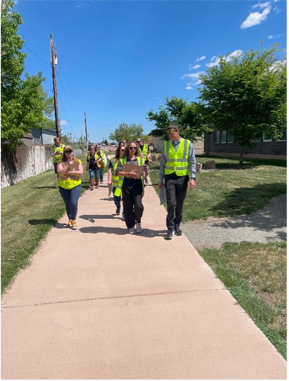 People in yellow safety vests walking on a sidewalk towards the camera. It is sunny.