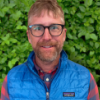 Man with glasses and a blue vest smiling in front of greenery 