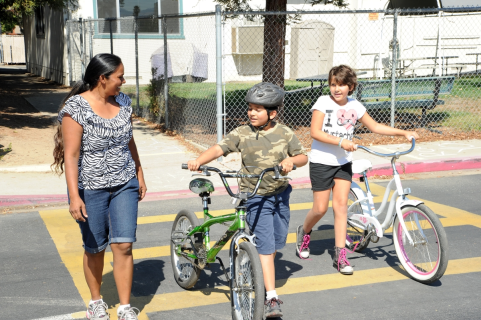 An adult with two children walk in a crosswalk with their bikes by their sides. 